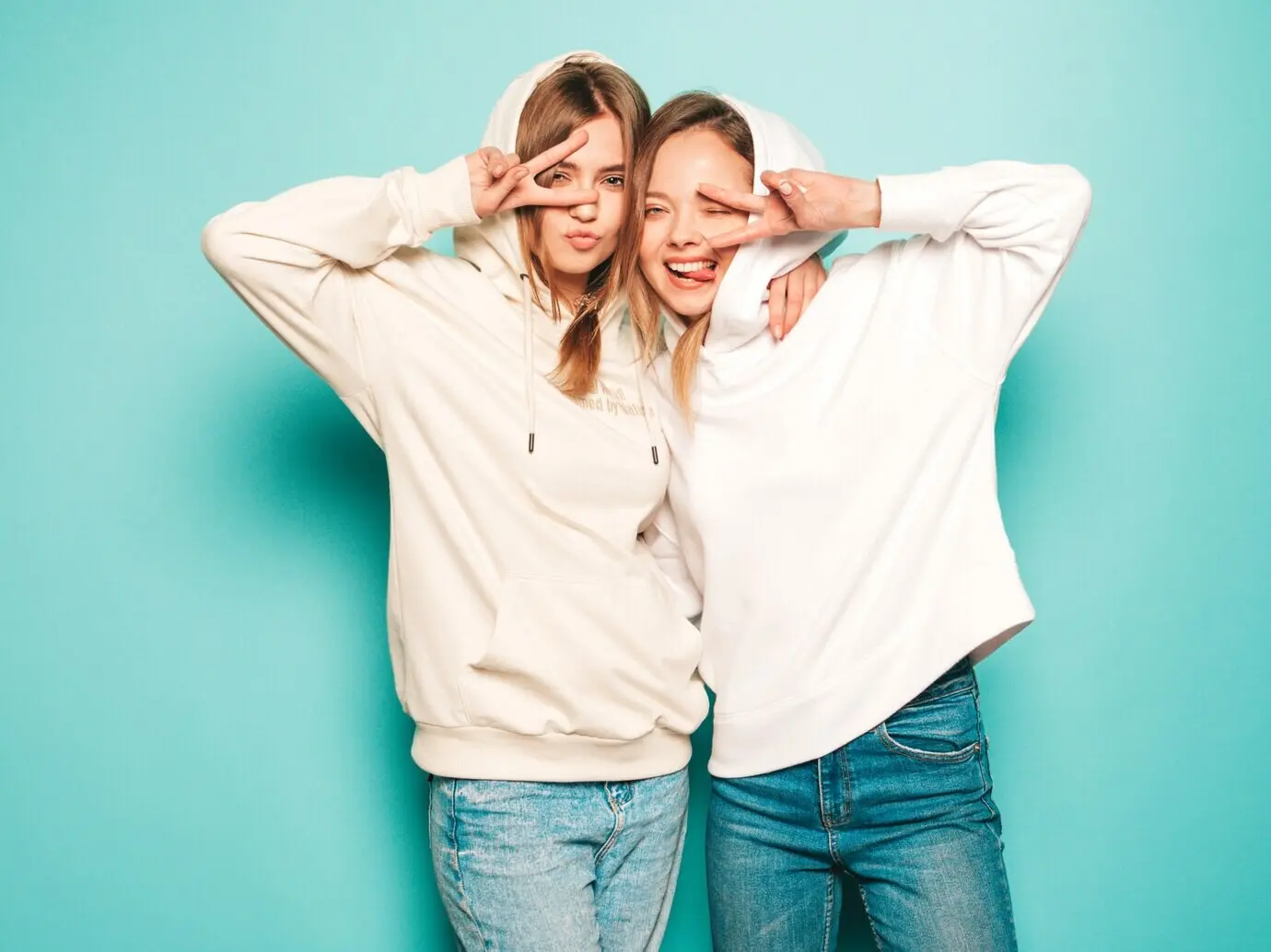 Zwei junge, schöne, blonde, lächelnde Hipster-Mädchen in trendigen sommerlichen Hoodie-Outfits. Sexy, unbeschwerte Frauen posieren nahe einer blauen Wand. Trendige und positive Models zeigen das Peace-Zeichen mit Sonnenbrillen.