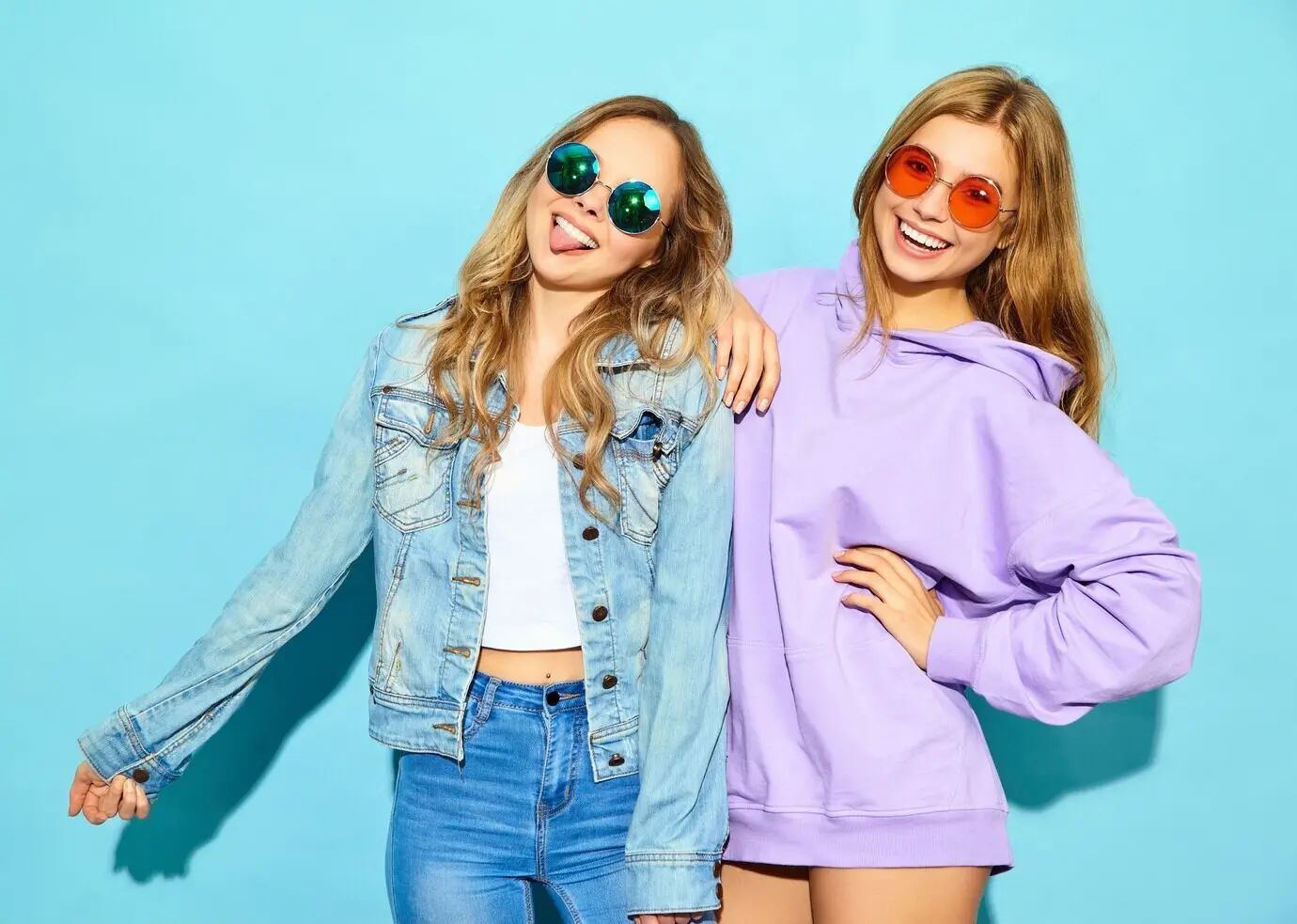 Zwei junge, schöne, blonde, lächelnde Hipster-Frauen in trendiger Sommerkleidung. Sexy, unbeschwerte Frauen posieren mit Sonnenbrillen nahe einer blauen Wand. Gut gelaunte Models flippen aus.